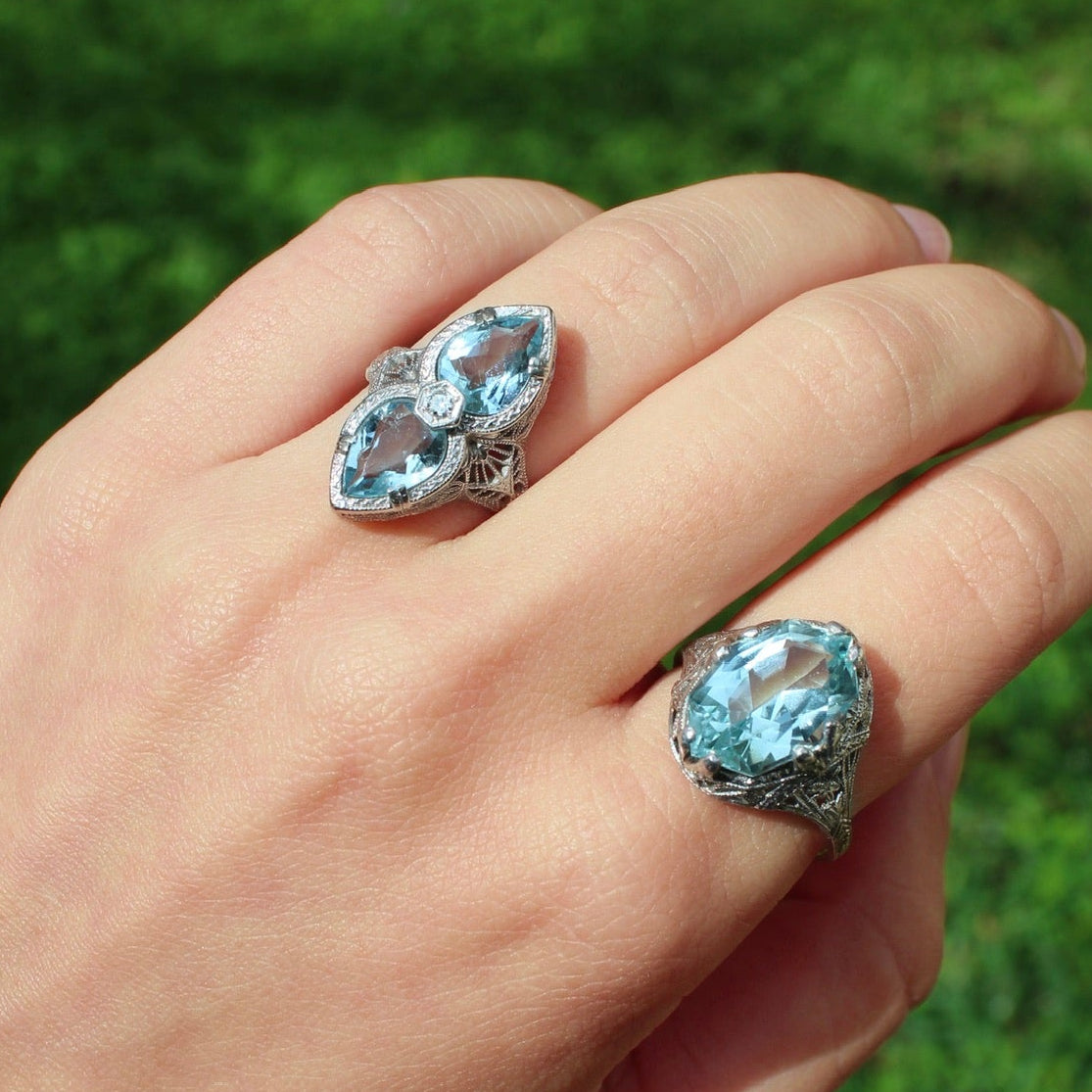 Hand wearing two rings with blue gemstones against a green grass background